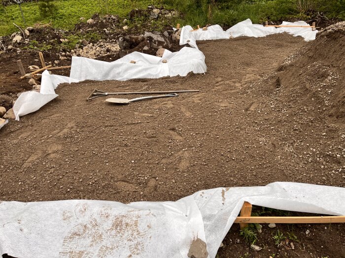 Markyta förberedd för växthusgrund, med synlig markduk i kanterna och verktyg på ytan. Omgiven av jord och stenar, klart för makadam.