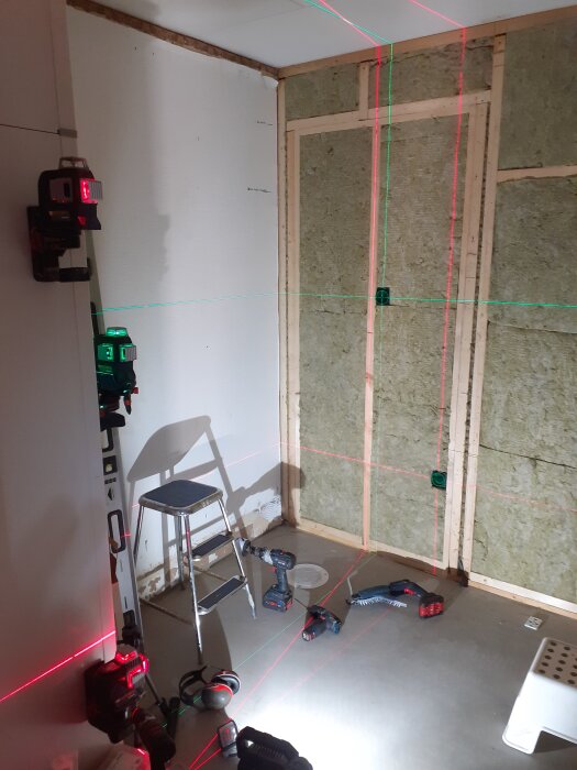 Byggprojekt med monterade skivor, stege, verktyg och lasermätare som projicerar röda och gröna linjer på vägg och golv i ett rum under renovering.