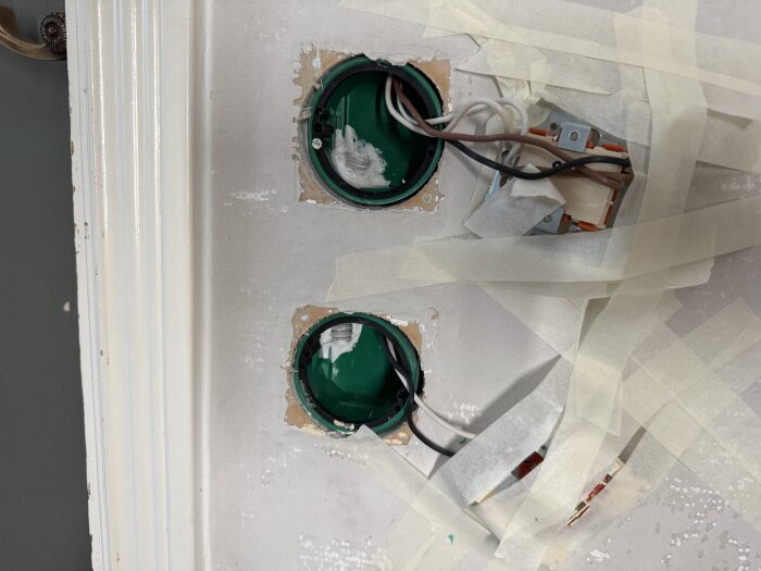 Elektrisk installation med två gröna eldosor monterade på en vägg, omgivna av täckande tejp. Sladdar och ledningar synliga.