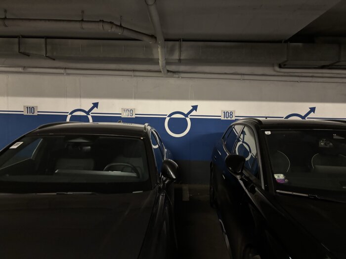 Två bilar parkerade i ett garage med blå och vita symboler på väggen, numrerade parkeringsplatser 108-110 syns mellan stolparna.