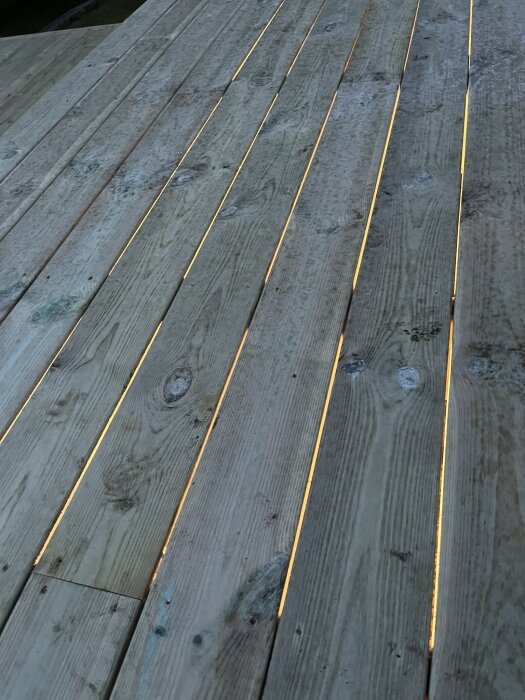 Trätrall med belysning under som skiner genom springorna, visar träets mönster och struktur på en altan i skymning.