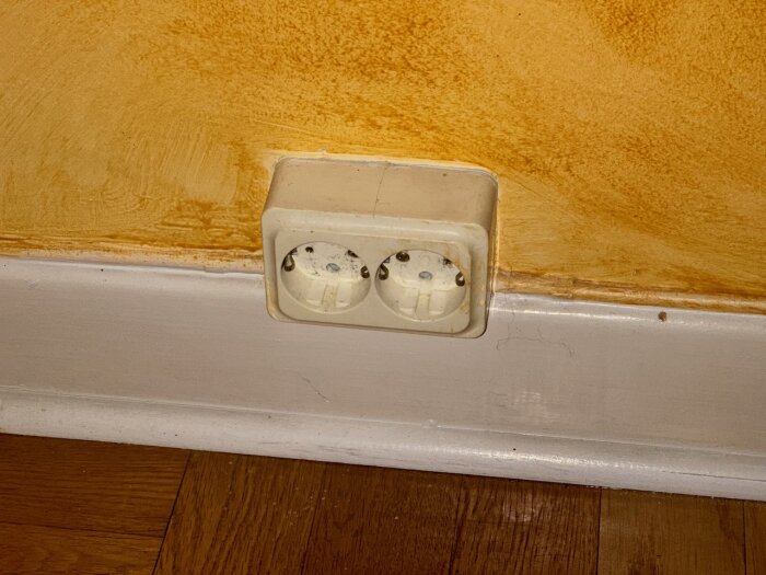 Dubbelt eluttag på vägg ovanför golvlist med gul bakgrund.