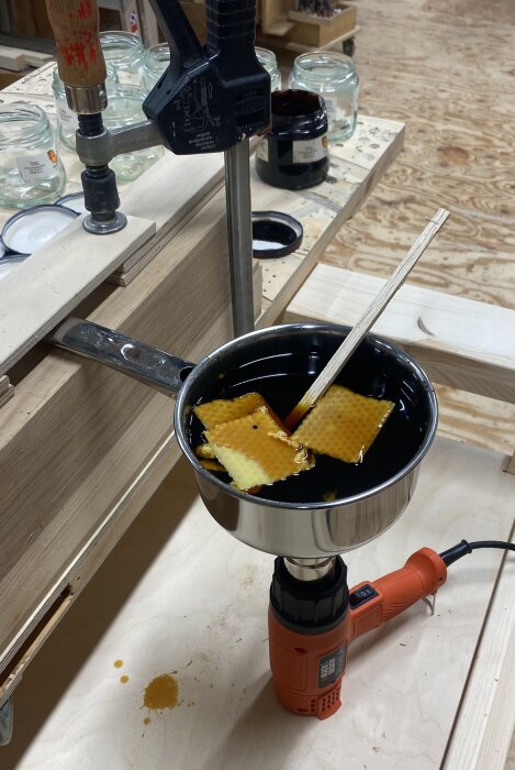 Metallkärl med bivax och tjära smälter över en varmluftspistol, omgiven av burkar på träbord.