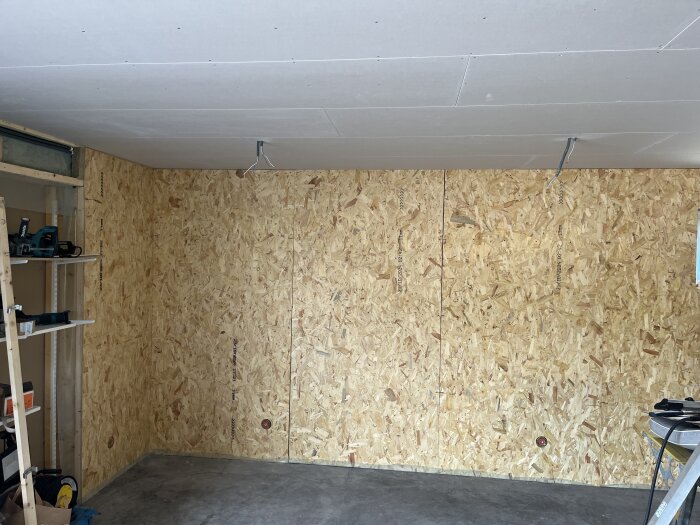 Väggar av OSB-skivor och ett gipstak i en pågående renovering, med lagerhyllor fyllda med verktyg på vänster sida.