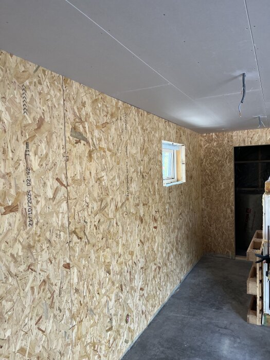 OSB-skivor på väggarna och gips i taket i ett rum under konstruktion, med ett fönster och synliga kablar i taket.