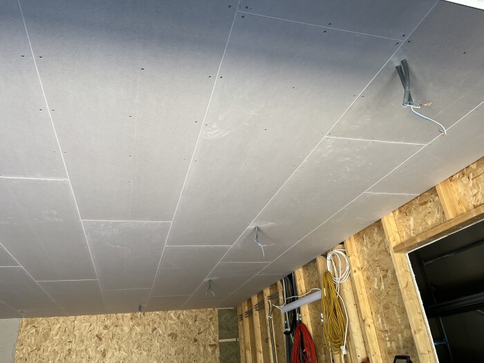 Gipstak med elkablar i ett rum under renovering, osb-skivor på väggarna, olika verktyg och kablar hänger på väggen.