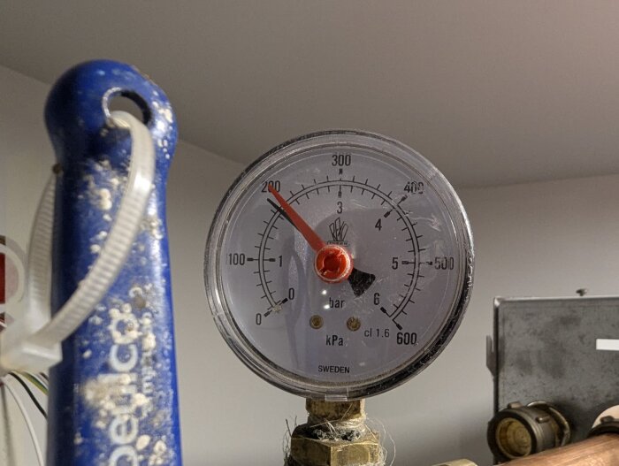 Manometer visar tryck på 1.95 bar i ett VVS-system, omgiven av blå rördel och metallkopplingar i ett inomhusutrymme.