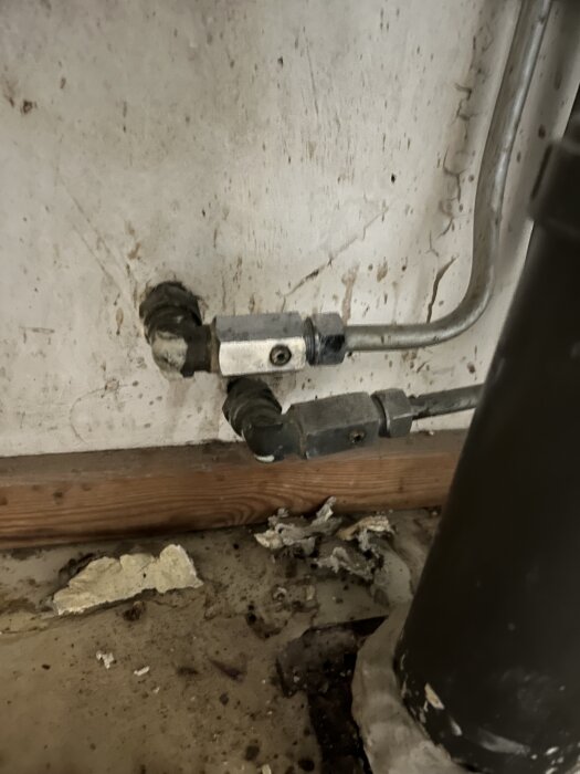 Rörkopplingar och en fastnad Ballofix-ventil med trasiga skruvspår på en vägg, tillsammans med rost och smuts på golvet.