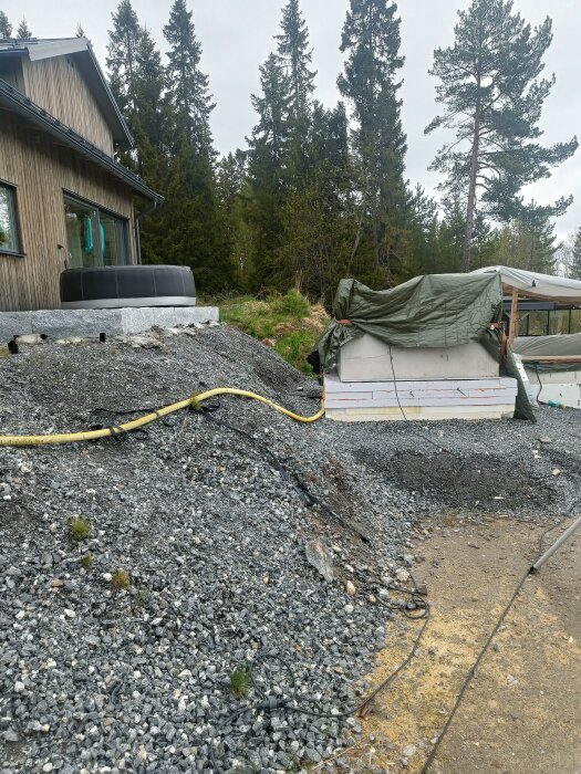 Grå slänt mellan hus och ett täckt område, troligtvis teknikrum. Slänten är brant och fylld med grus, en gul slang löper tvärs över.