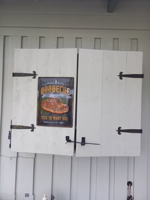 Vita träskåpdörrar med svarta gångjärn, delvis öppnade. Dörrarna har slagit sig och en BBQ-skylt är fastsatt på dem.