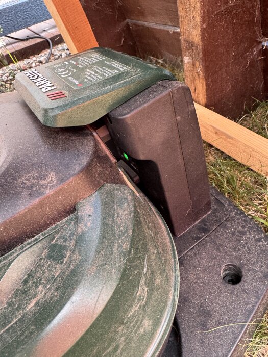 Grön och svart gräsklippare på laddningsstation med märkning "Parkside", inställd mot trävägg, står på gräs och grus.