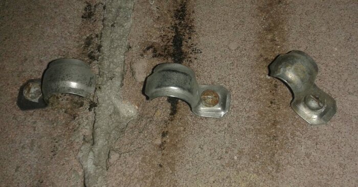 Tre metallklämmor fastskruvade på en sliten betongyta med sprickor, troligtvis från ett gammalt sågverk.