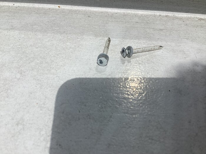 Två metallskruvar med gängor ligger på en jämn yta i solljus.