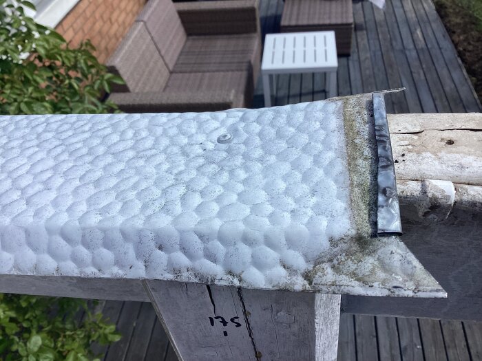 Plåt ovanpå träbalk med spik med gängor fastsatt, byggmaterial synligt på terrass med möbler i bakgrunden.