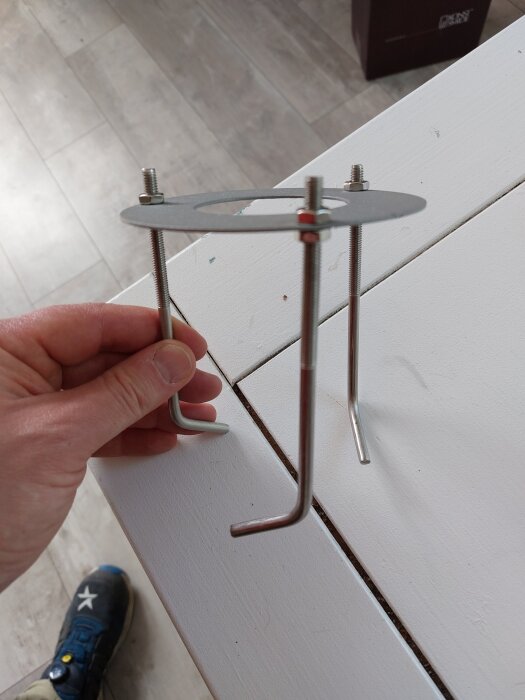 Hand håller ett metallfäste med tre stänger och muttrar, som används för att installera pollare vid markstensläggning.