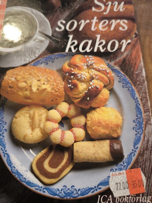 Omslag av en kokbok med flera olika kakor, inklusive kanelbulle, syltkaka, chokladöverdraget bakverk och sockrad krans, på en blåvit tallrik.