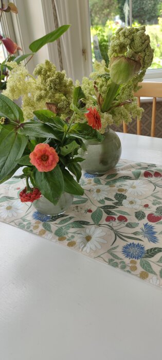 Blommor i en vas på ett bord med en duk som har blommönster. Soligt fönster i bakgrunden.