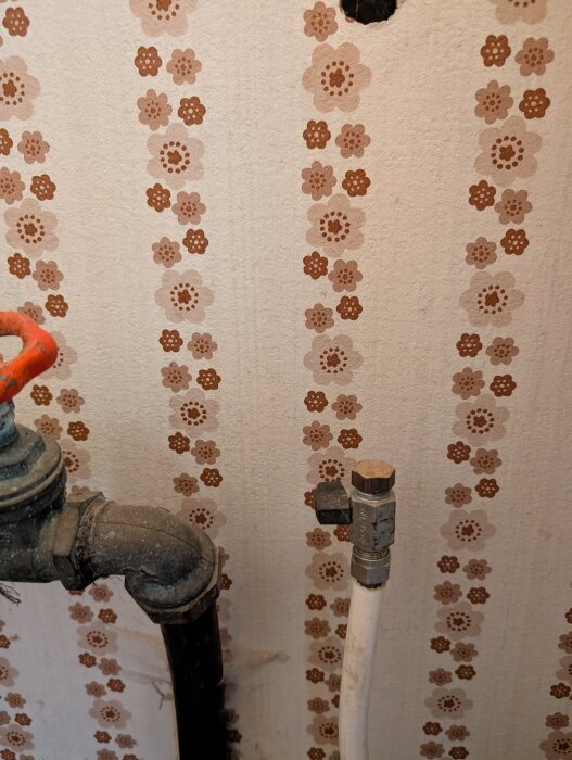 Vägg med blommigt mönster och två rör, ett metallrör med en kranventil och ett vitt plast- eller metallrör med anslutning.