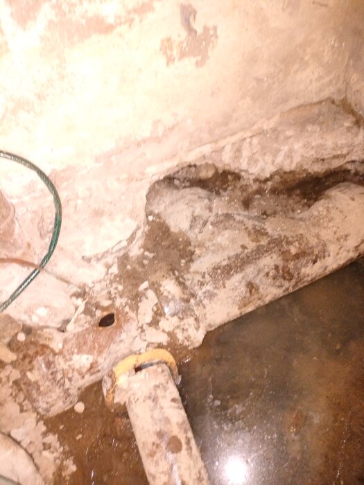Gammal avloppsledning med sprucken omgivning och vattenläckage på betonggolv i källare.