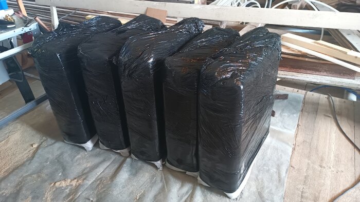 Svart plastinpackade föremål på ett golv i ett byggutrymme, med olika verktyg och material i bakgrunden.