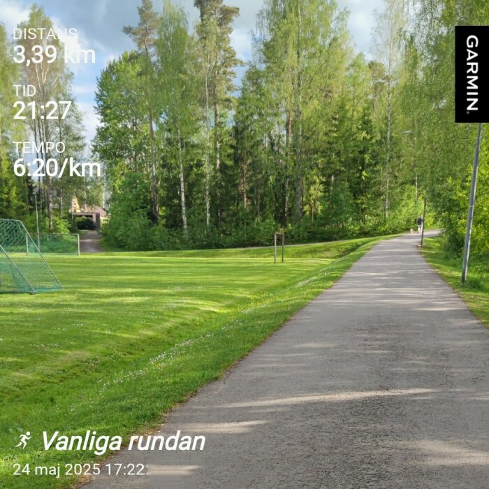 Gångväg genom grön park med fotbollsmål till vänster, omgiven av träd under en klar himmel, text med löpdistans och tid från Garmin.