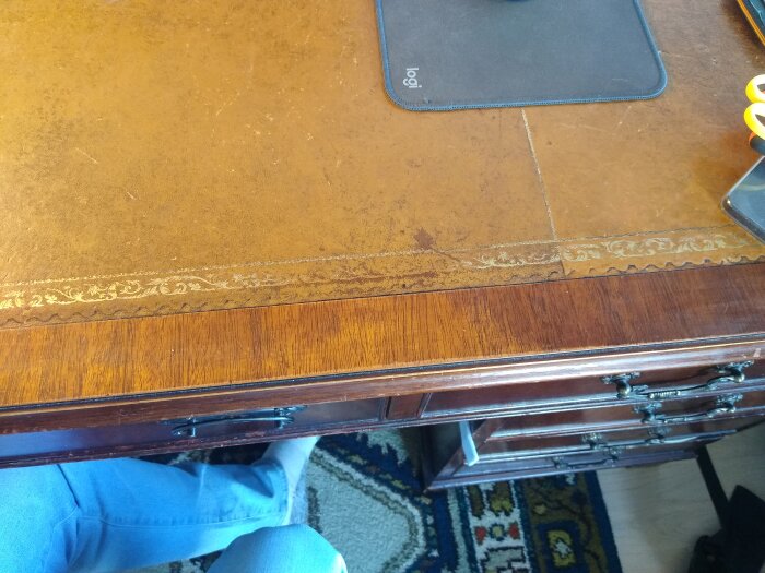 Engelskt skrivbord med synligt slitage och avskavd lack på fanér, särskilt där högerarmen vilar. Detaljerade dekorativa element längs kanten.