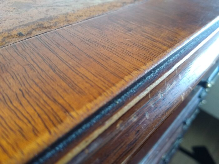 Närbild av fanérkant på engelskt skrivbord med slitage, lacken har slitits från mörk brun till ljus brun och orange, viss del matt utan glans.