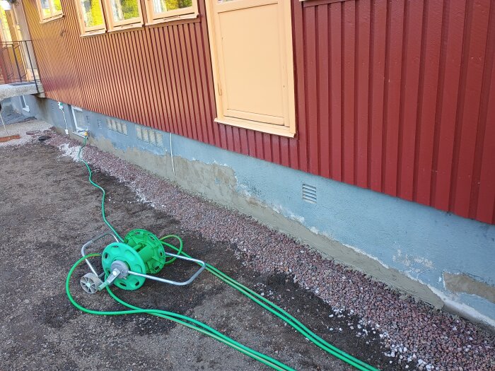 Fasad med lagat puts och grön trädgårdsslang på marken framför ett hus med röd träpanel och gul dörr.