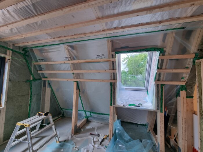 Byggnad under renovering med regelväggar och plastöverdrag. Ett fönster monterat i taket, verktyg och material synliga på platsen.