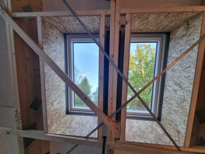 Byggprojekt med två fönster omgivna av trä- och skivkonstruktion, sedd inifrån.