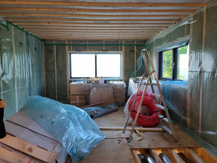 Byggprojekt inomhus med väggar täckta av plast, stege, rör och byggmaterial på golvet i ett rum med stora fönster.