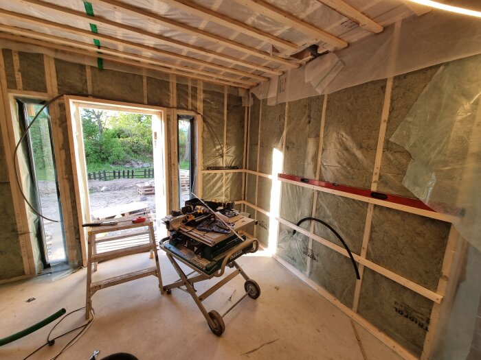 Rum under renovering med reglar och plastisolering på väggar, arbetsbänk med verktyg och material, öppning mot utsidan med grönt landskap.