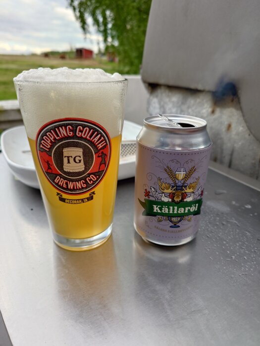 Ölglas med "Toppling Goliath" logotyp bredvid en burk "Källaröl" på metallbänk utomhus.