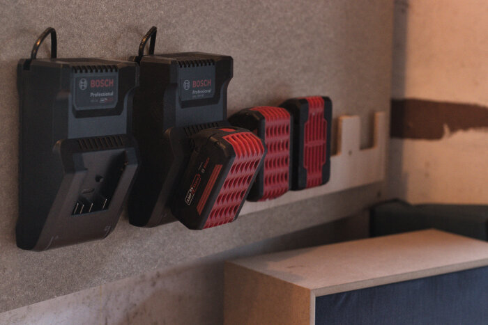 Väggmonterad laddstation med två Bosch laddare och fyra batterier, monterade på plywood i ett garage.
