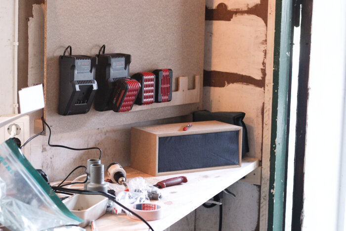 Väggmonterade laddare och hållare för 4 batterier i ett garage, med en hylla nedanför fylld med verktyg och material.