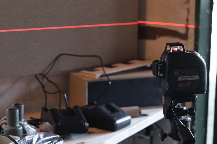 Bosch laserverktyg projicerar en röd linje på väggen, omgivna av laddare och byggmaterial i en verkstadsmiljö.