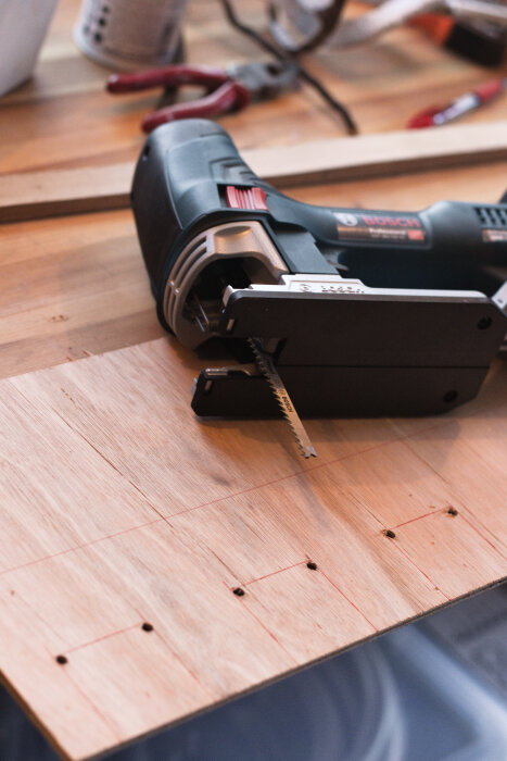 Sticksåg från Bosch används för att såga ut former i en plywoodskiva, placerad på en arbetsbänk, med märkningar för hål och linjer.