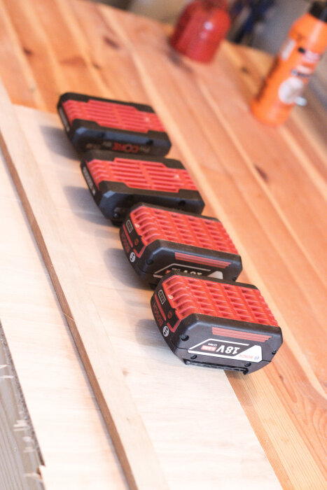 Fyra röda och svarta Bosch 18V batterier placerade på en träyta i ett garage.