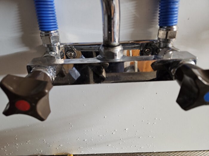 Två kranar monterade på en vägg, anslutna med flexibla blå slangar och halvkopplingar.