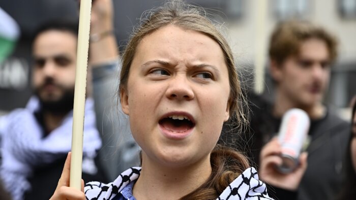 Ung person vid protest, håller en skylt, klädd i svartvit sjal, med andra demonstranter i bakgrunden.