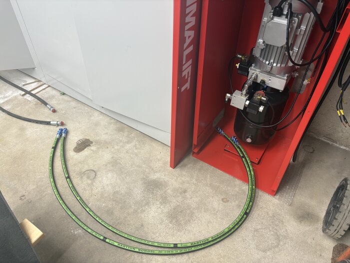 Gröna hydraulslangar med kopplingar ligger på ett betonggolv, anslutna till en röd maskin med motor och pump. En bit trä och fotavtryck syns på golvet.