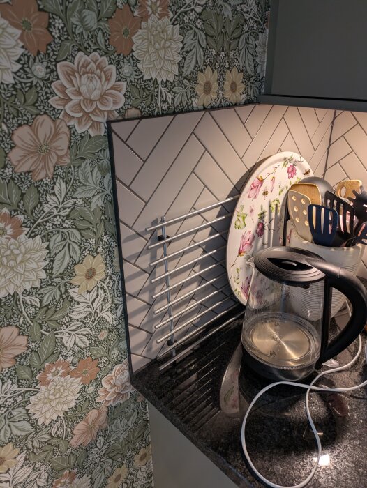 Hörn i kök med blommig tapet och fiskbensmönstrat kakel. Svart bänkskiva med glasvattenkokare och köksredskap i en behållare.