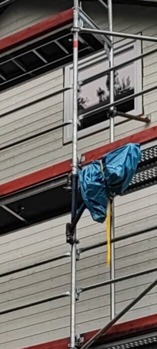 Byggställning med plåtfasad i bakgrunden och en insvept elvinsch på sidan skyddad av en blå sopsäck.