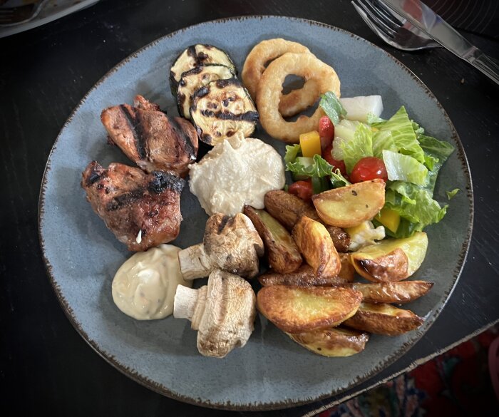 Grillad flÀskfilé, kamben, champinjoner, squash, ugnsstekt potatis, sallad, hummus och bearnaisesÄs pÄ en tallrik.