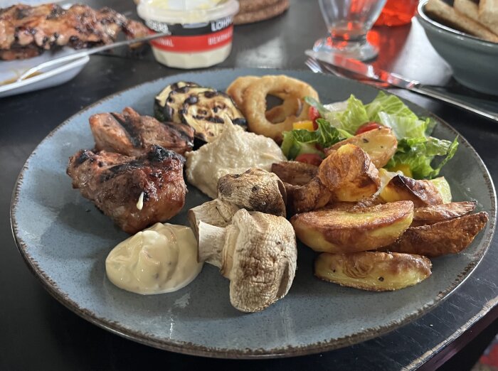 Grilltallrik med flÀskfilé, champinjoner, squash, ugnspotatis, lökring, sallad, hummus och bearnaisesÄs pÄ en mörk tallrik.