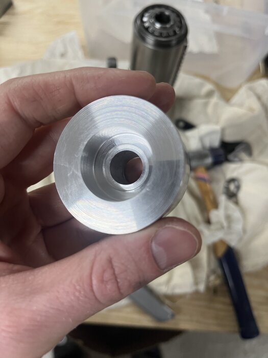 Hand som håller en svarvad aluminiumdel, en konisk cylinder, ovanför en bänk med verktyg i bakgrunden.