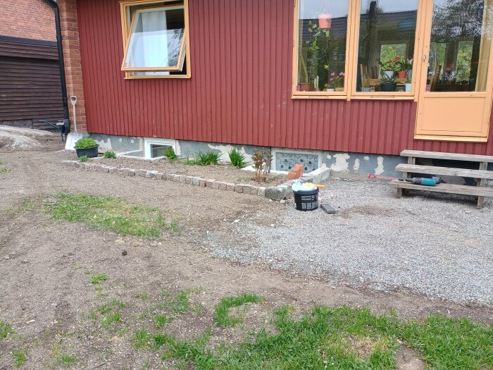 Fasad på baksida av hus med grundmålad vägg, krukväxter under fönster, trappa och grusad yta.