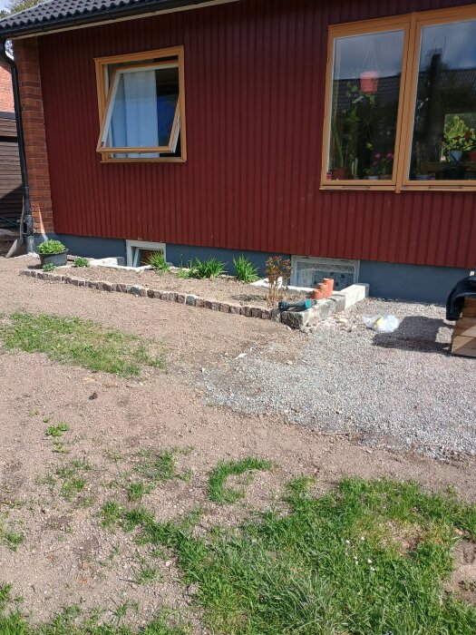 Bakgård med röd husvägg, delvis grundmålad. Grus och nyplanterad rabatt framför väggen.