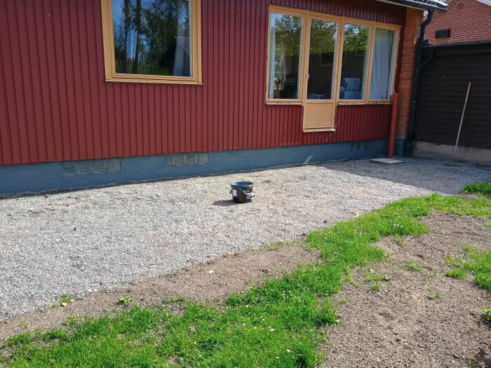 Baksidan av ett hus med röd fasad och nygrundmålat område nära gräsmatta och grusgång. En buckett och pensel ses på marken.