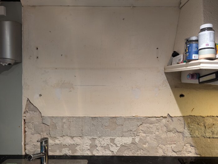 Nedre delen av köksvägg med gammal puts och spår av borttaget kakel ovanför en diskbänk, samt öppna hyllor med olika burkar ovanför.